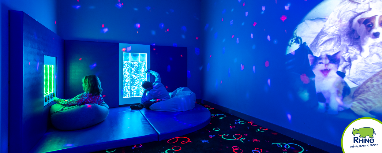 Two children sit on beanbags inside a sensory dark room, interacting with light up wall panels on the walls of the room