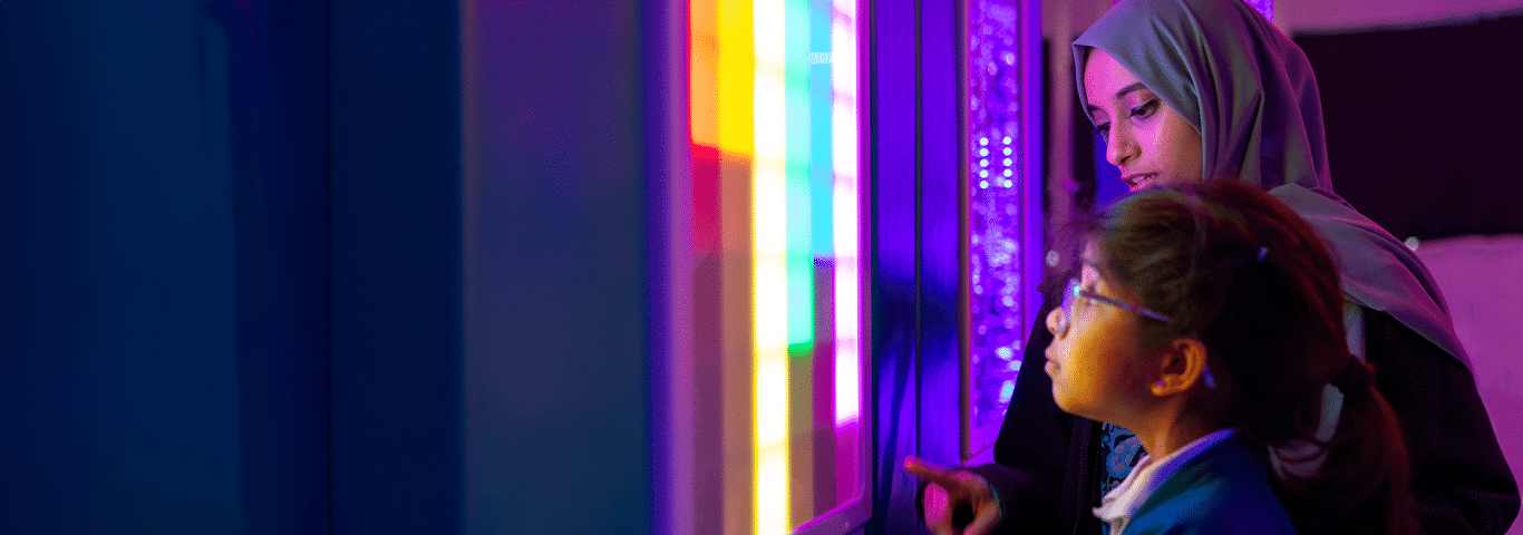 A young girl and her mother engaging with a colorful light panel featuring interactive buttons in a sensory room.