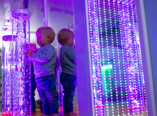 A toddler smiles as they stand next to a bubble tube