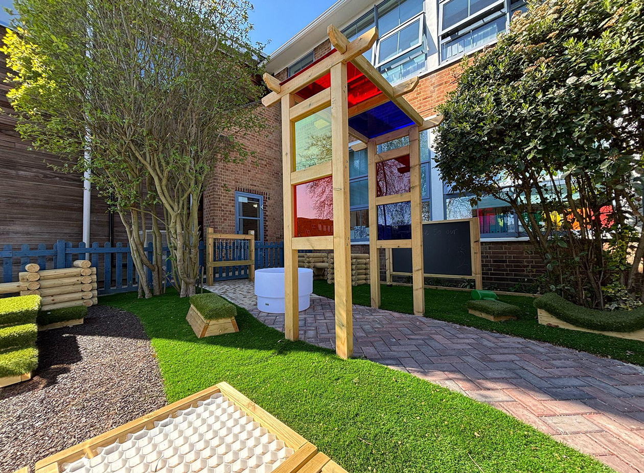 An outdoor sensory garden with a colourful wooden play tower, artificial grass, tactile sand trays, raised planting beds, and paved pathways surrounded by natural shrubs and trees